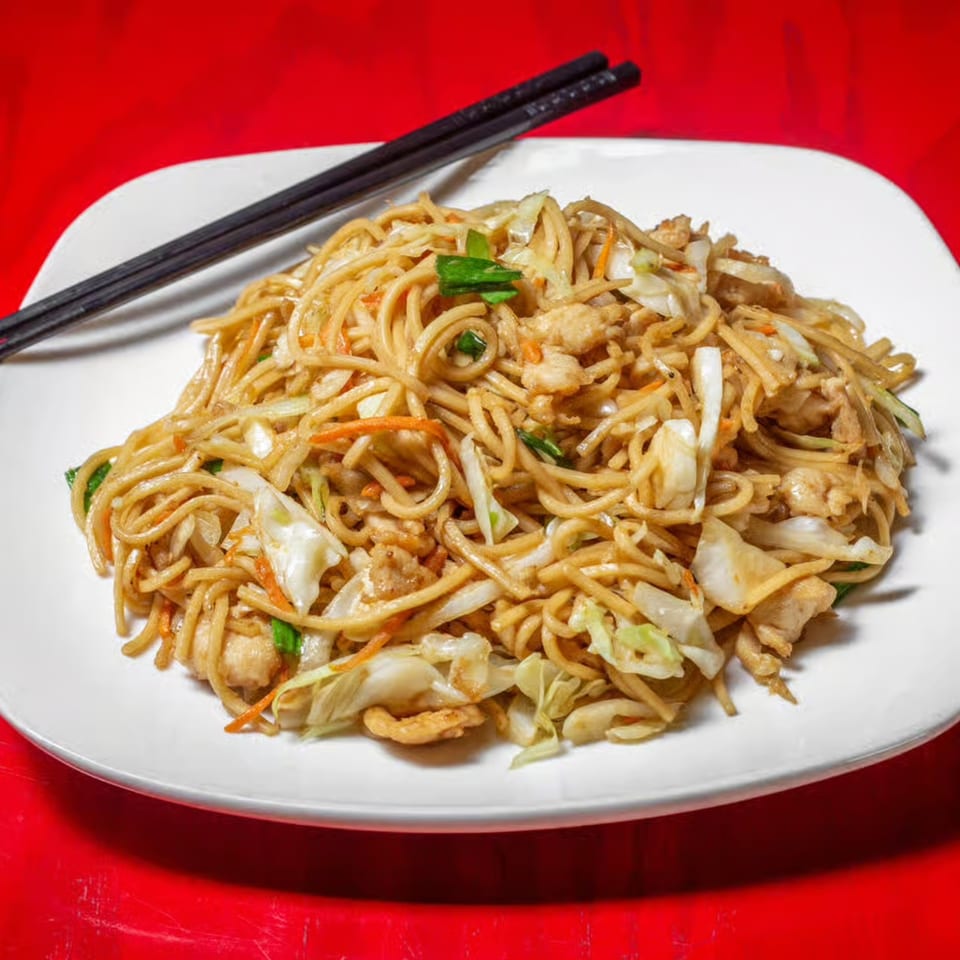 A white plate of Asian noodles with chicken, cabbage, carrots, and green onions, with chopsticks.