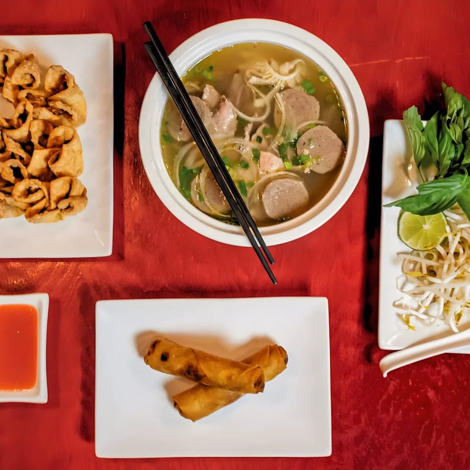 Overhead view of Heavenly Asian Cuisine's pho, crispy wontons, spring rolls, and fresh herbs on a red table.