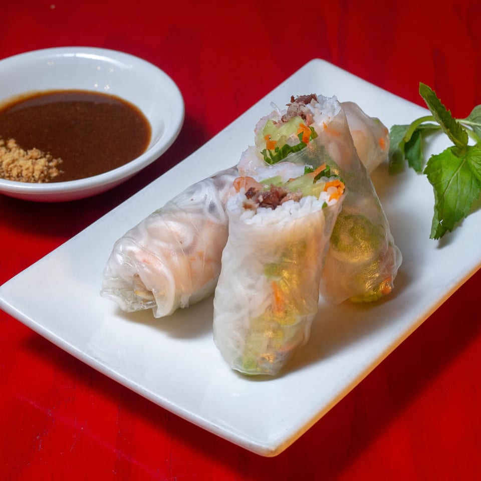 Three fresh spring rolls with vegetables and meat, served with a side of peanut sauce and mint.