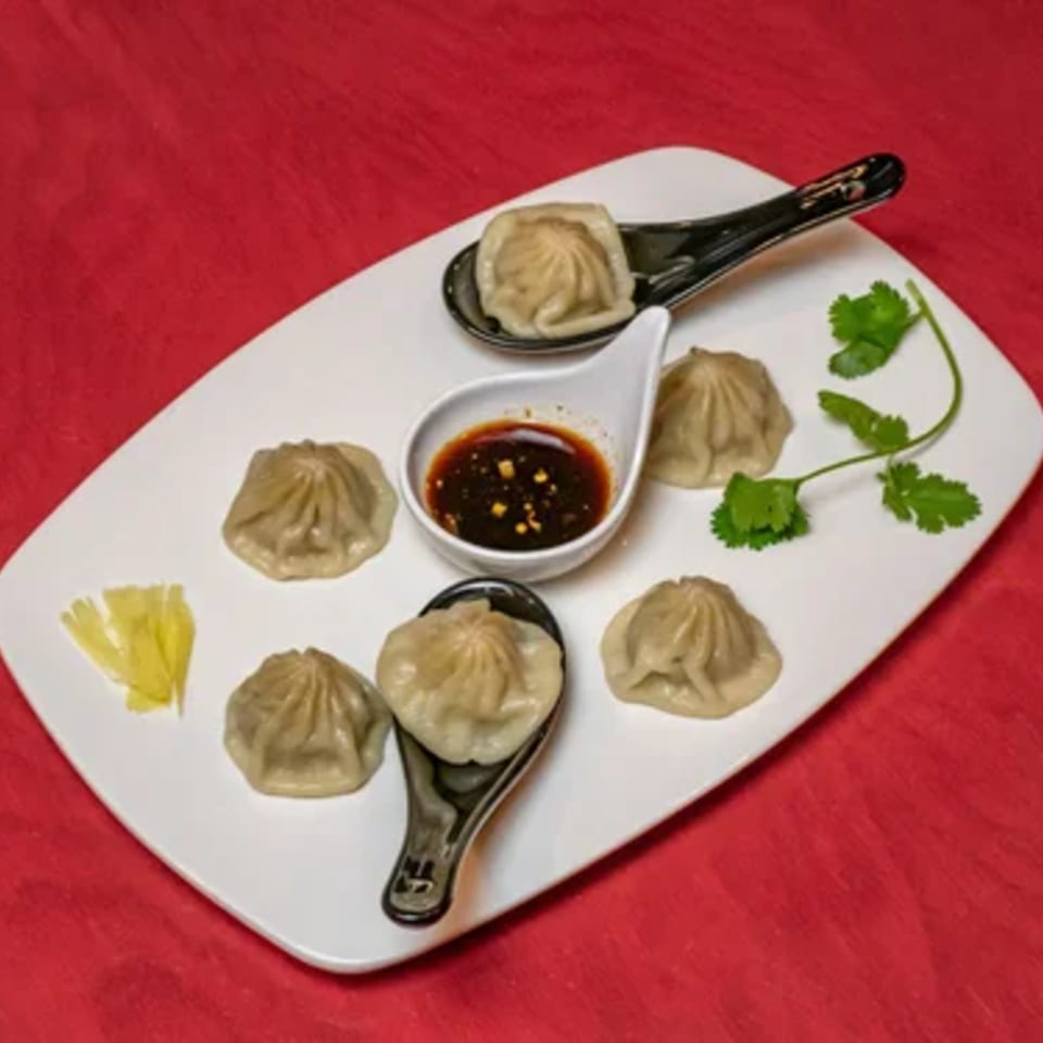 A white plate with six Asian dumplings, a small bowl of dipping sauce, and cilantro on a red background.