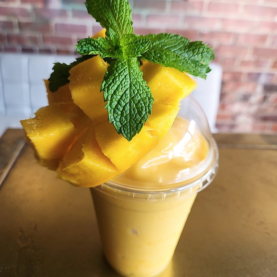 A mango smoothie in a clear cup, garnished with fresh mango slices and mint leaves.